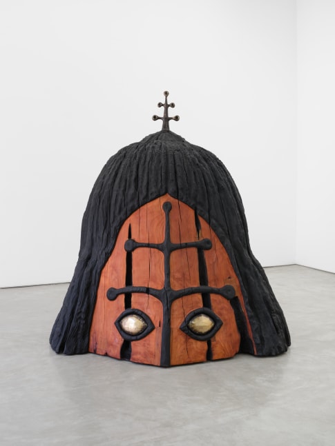 A redwood sculpture, a large top portion of a face. Black symmetrical design on the center, with aluminum eyes. And a metal antenna figure peeking out from the top of the head.