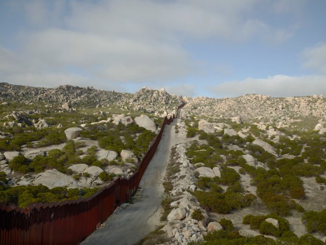 A photograph by Richard Misrach titled 'Wall, Tierra Del Sol, California', 2015 featuring a rocky landscape and the border wall.