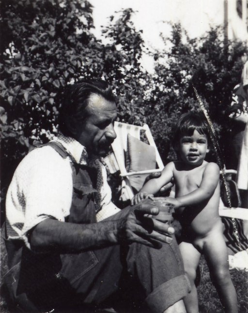 Man in overalls and a white collared shirt sitting on the grass with a female toddler standing beside his left knee