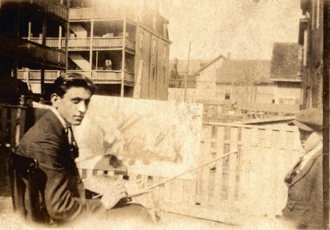 Faded sepia photograph of a seated young man holding a palette, brushes, and other artist materials while painting a landscape al-fresco in a residential neighborhood