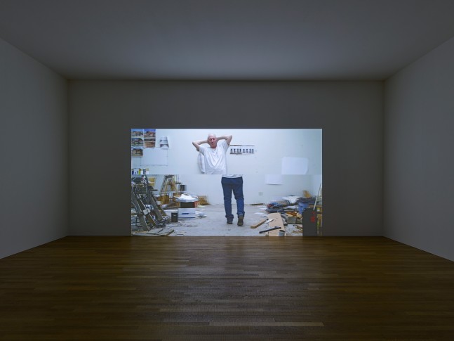 installation view of a darkened gallery with a projected image of a man walking with his hands behind his head in a contrapposto pose