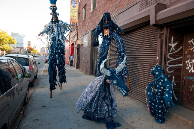 Photograph of performance on the street in Brooklyn, New York featuring several stilt-walkers and people wearing indigo blue costumes