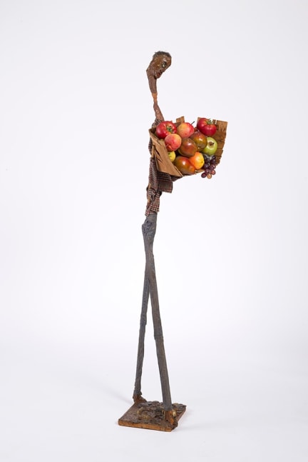 A sculpture made out of Hogmawg, fabric, and mixed media that depicts a tall man carrying a basket filled with fruit such as apples, grapes and even tomatoes.