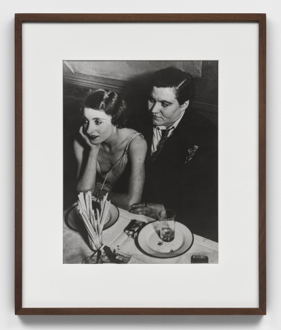 A black and white photographic print by Brassai of a couple at a white top table with plates, glasses and a pack of cigarettes