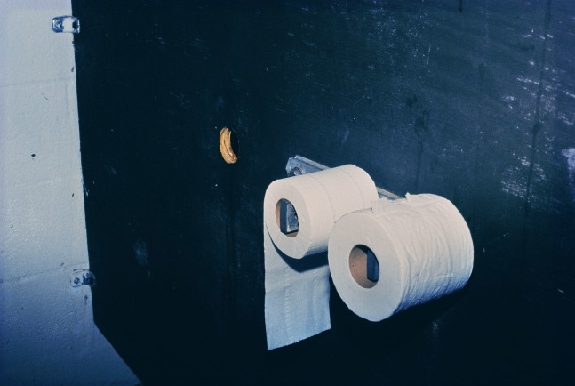 Image of a bathroom stall with two rolls of toilet paper and a cut out hole through the wall by Mickey Aloisio