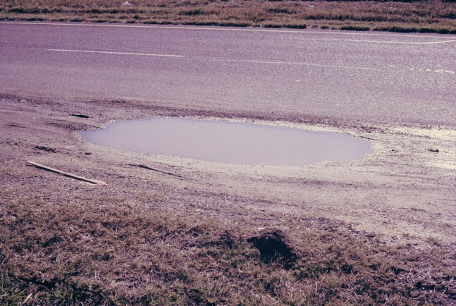 Image of a puddle surrounded by dirt on the side of a road by Mickey Aloisio