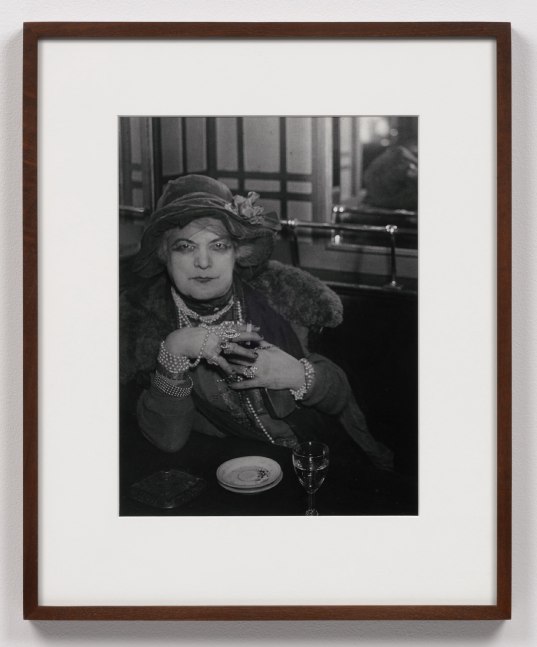 A black and white photographic print by Brassai depicting Bijou holding a cigarette, wearing different types of jewelry at the Bar de la Lune