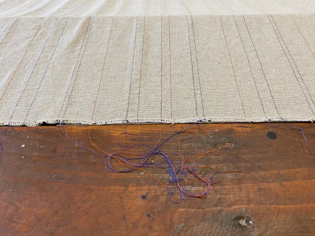 Color photograph of a textile based artwork comprised of a large rectangular piece of tan linen draped over a trap door opening in the floor. Artwork by Gabriela Salazar, installed at the Al Held Foundation, 2021. Exhibition curated by Olga Dekalo and River Valley Arts Collective.
