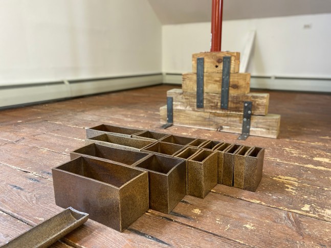 Color photograph of a ceramic artwork by Gabriela Salazar, installed at the Al Held Foundation, 2021. Comprised of small rectangular vessels arranged in a pyramid shape on the floor. Exhibition curated by Olga Dekalo and River Valley Arts Collective.