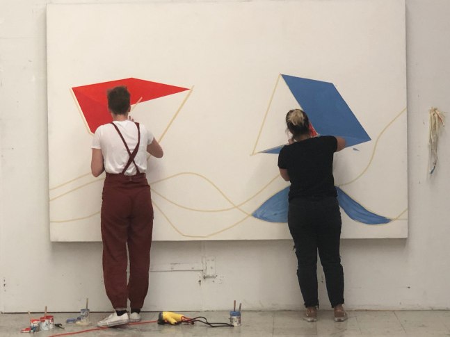 Two students create geometric shapes with masking tape across the surface of a prepared canvas.