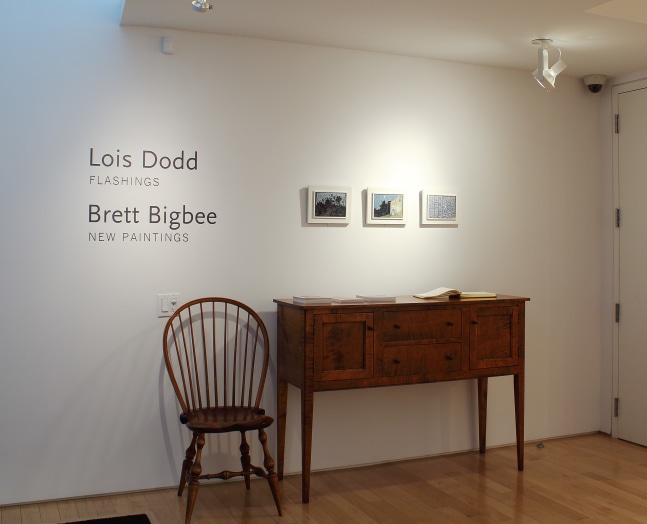 Photograph showing a front view of an exhibition space, with three small paintings hanging on a white wall above a table and chair