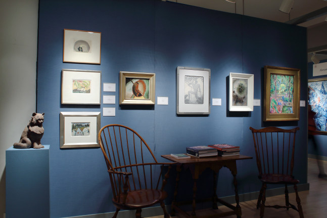 Photograph showing a view of an exhibition booth, with seven paintings hanging on a blue wall and a sculpture on a pedestal beside