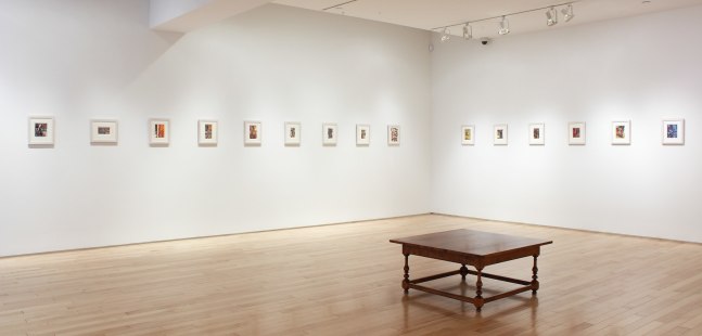 Photograph showing a corner view of an exhibition space, with fifteen small framed paintings hanging on white walls