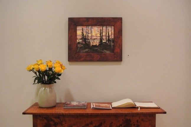 Photograph showing a view of an exhibition space, with a small framed painting hanging over a table with a guestbook and vase of flowers on it