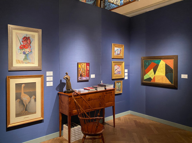 Photograph showing a corner view of an exhibition with seven paintings in various styles and sizes hanging on dark blue walls