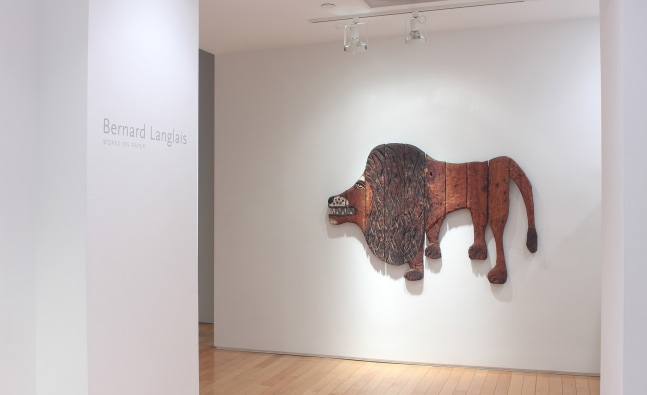 Photograph of a view of an exhibition space, with a wooden sculpture hanging on a white wall and text that reads &quot;Bernard Langlais: Works on Paper&quot; on a wall to the right