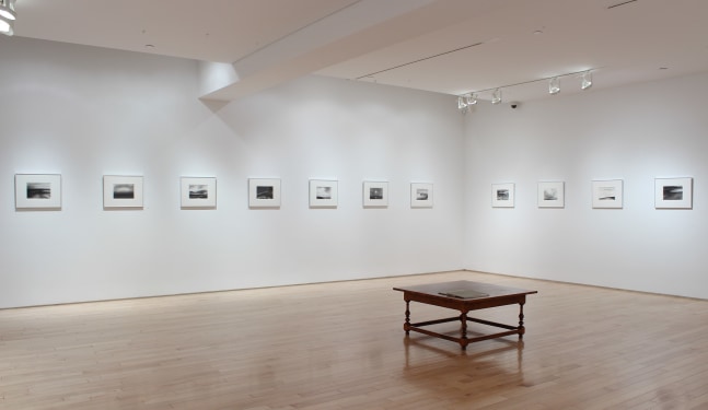 Photograph showing a corner view of an exhibition space, with thirteen drawings hanging on white walls