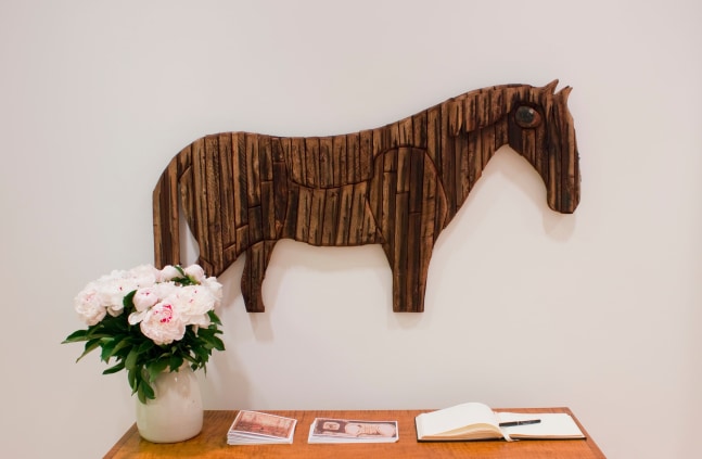 Photograph showing a view of an exhibition space, with a wood sculpture of a horse hanging over a table with a vase of flowers on it
