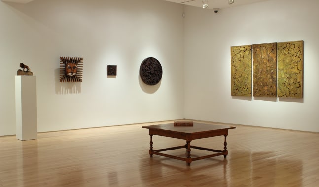 Photograph showing a corner view of an exhibition space, with five sculptures hanging on white walls