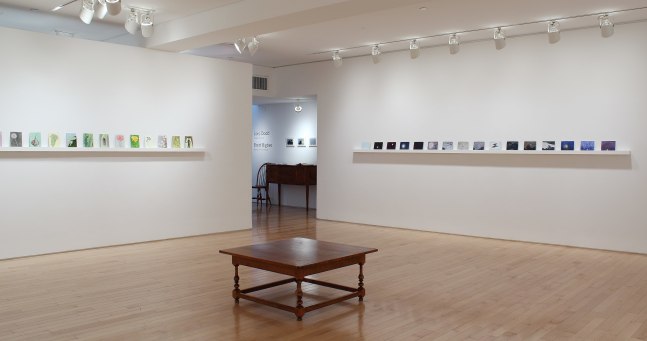 Photograph showing a corner view of an exhibition space, each wall holding dozens of paintings on a long shelf