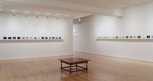 Photograph showing a corner view of an exhibition space, each wall holding dozens of small paintings on a long shelf