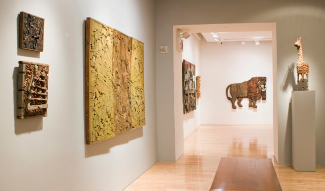 Photograph showing a view of an exhibition space, with six sculptures hanging on the walls and one sculpture on a pedestal to the right