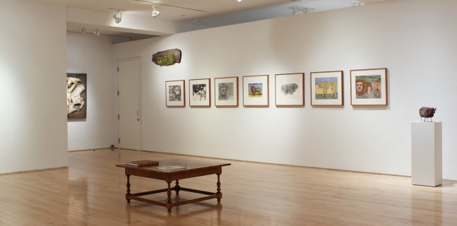 Photograph showing a corner view of an exhibition space, with seven framed drawings hanging on a white wall, and a small sculpture on a pedestal to the right