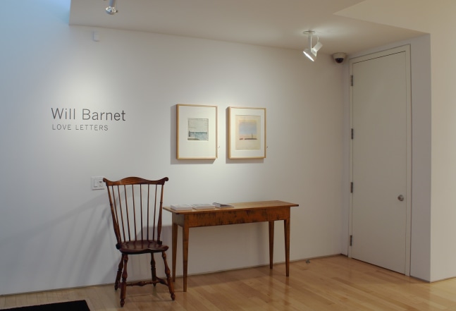 Photograph showing a front view of an exhibition space, with two paintings hanging on a white wall above a table and chair