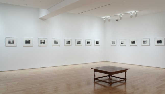 Photograph showing a corner view of an exhibition space, with fourteen framed drawings hanging on white walls
