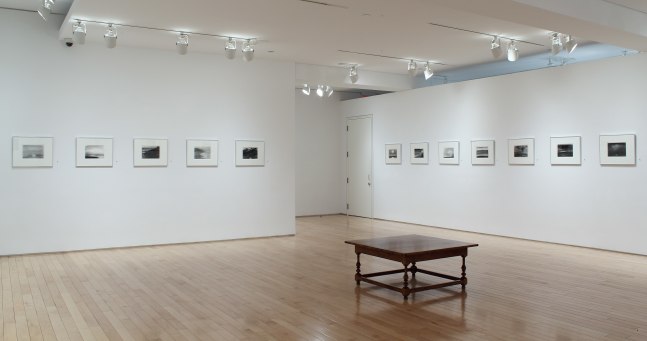 Photograph showing a corner view of an exhibition space, with twelve framed drawings hanging on white walls