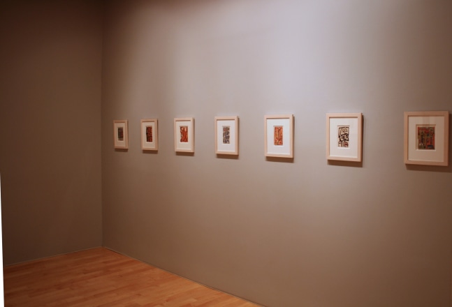 Photograph showing a corner view of an exhibition space, with seven framed paintings hanging on a grey wall