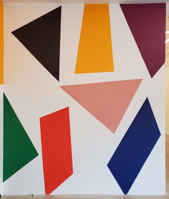 Photograph showing an abstract mural painted on a wall, with geometric shapes in black, yellow, pink, red, green, and blue arranged on a white wall