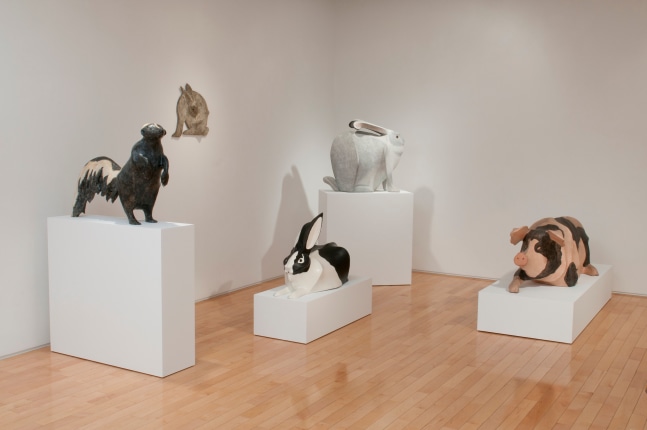 Photograph showing a corner view of an exhibition space, with four sculptures on pedestals and one hanging on the wall