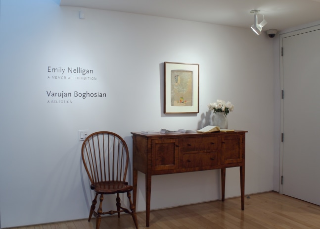 Photograph showing a front view of an exhibition space, with one painting hanging on a white wall above a table and chair