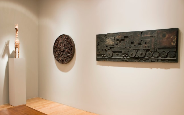 Photograph showing a view of an exhibition space, with two sculptures hanging on a wall and a sculpture on a pedestal to the left