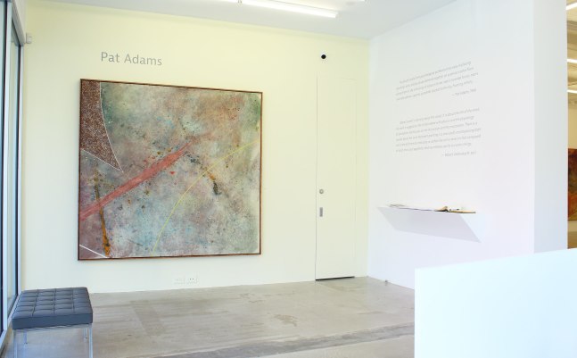 Photograph showing a corner view of an exhibition space, with one painting hung on a yellow wall and a white wall with text related to the exhibition