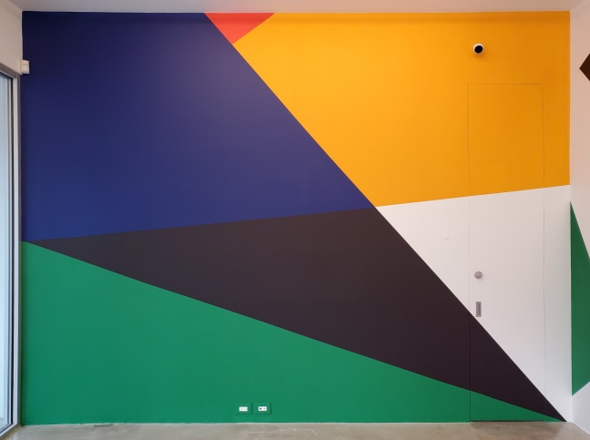 Photograph showing a geometric abstract mural painted on a wall, with interlocking shapes of green, black, blue, yellow, white, and red
