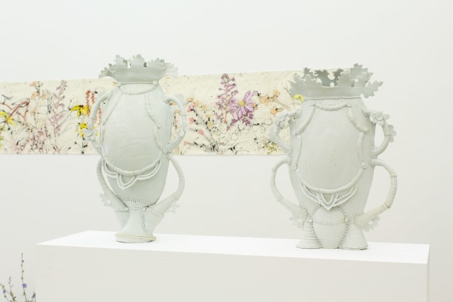 Photograph showing a detail of an exhibition, with two white porcelain sculptures in the foreground and a drawing on the wall in the background.