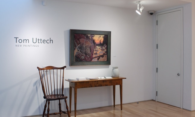 Photograph showing a view of an exhibition space, with a painting hanging on a white wall above a table and chair