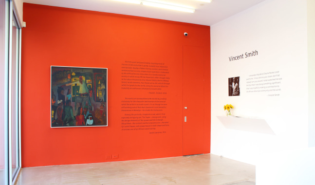 Photograph of the front corner of an exhibition space, showing a painting on a red wall with wall text beside, and a white wall with wall text