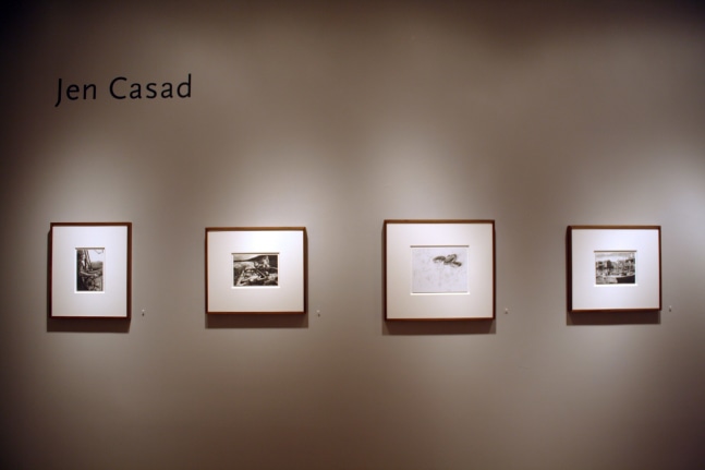 Photograph showing a view of an exhibition, with four small framed drawings hanging on a grey wall, and text that reads &quot;Jen Casad&quot; on the top left