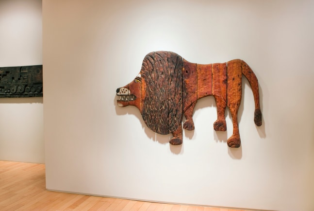 Photograph showing a view of an exhibition space, with a sculpture of a lion hanging on a white wall