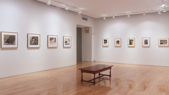 Photograph showing a corner view of an exhibition space, with nine framed paintings hanging on white walls