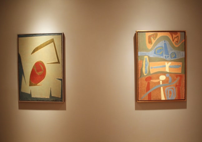 Photograph showing a view of an exhibition, with two framed paintings hanging on a grey wall