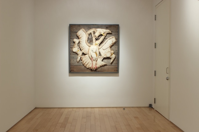 Photograph showing a view of an exhibition space, with a sculpture hanging on a white wall