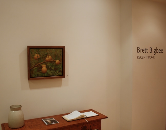 Photograph showing a view of an exhibition, with a painting hanging over a wood table and wall text that reads &quot;Brett Bigbee: Recent Work&quot; on the wall to the right