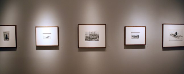 Photograph showing a view of an exhibition, with five small framed drawings hanging on a grey wall
