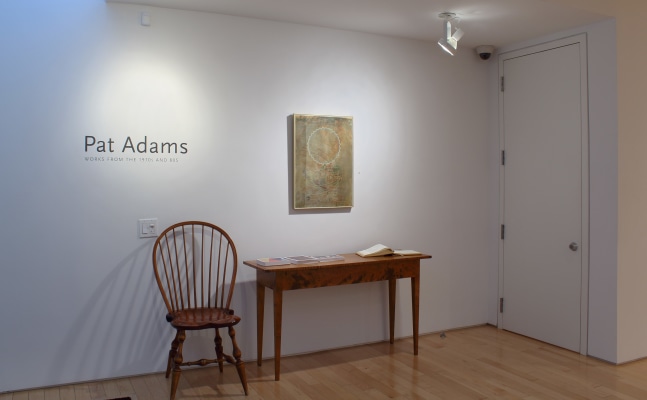 Photograph showing the front view of an exhibition space, with a painting hung on a white wall, and a table and chair below