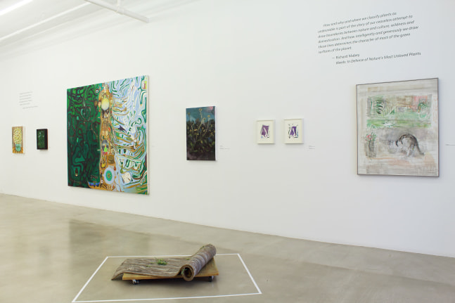 Photograph of a detail of an exhibition with seven paintings hung on a white wall and a sculpture on the ground in front