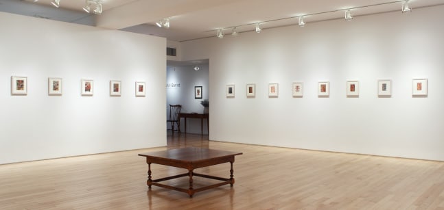Photograph showing a corner view of an exhibition space, with thirteen small framed paintings hanging on white walls